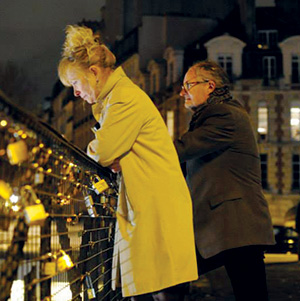 A woman in a light-colored coat and a man in a dark coat lean over a bridge covered with love locks at night, illuminated by nearby lights, with buildings in the background.