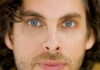 A close-up portrait of a man with light blue eyes, wavy brown hair, and a short beard, looking directly at the camera. He wears a jacket and a shirt, with a neutral background.