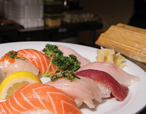 A white plate with assorted sushi nigiri, including salmon, tuna, and white fish, garnished with parsley, a lemon slice, and pickled ginger. A bamboo mat is visible in the background.