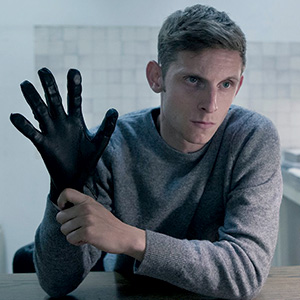 A serious young man in a gray sweater puts on a black glove while sitting at a table in a room with white tiled walls.
