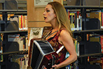 A woman with long blonde hair plays an accordion and sings in front of bookshelves in a library.