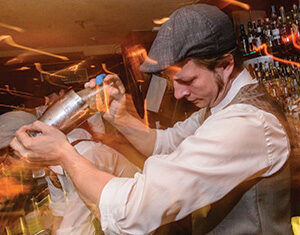 A bartender wearing a cap and vest vigorously shakes a cocktail shaker behind a dimly lit bar, with blurred lights and bottles in the background.