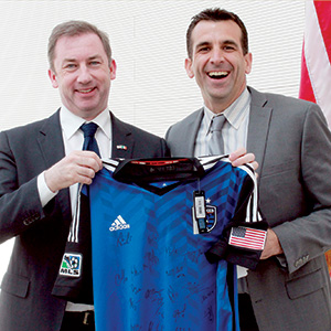 Two men in suits smile while holding up a blue soccer jersey with signatures. One man has an American flag pin on his lapel, and an American flag is visible in the background.