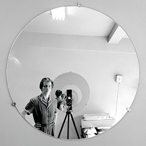 A black-and-white photo shows a person in vintage clothing taking a self-portrait with a camera on a tripod, reflected in a round wall mirror. The room appears simple, with a light fixture and some equipment visible.