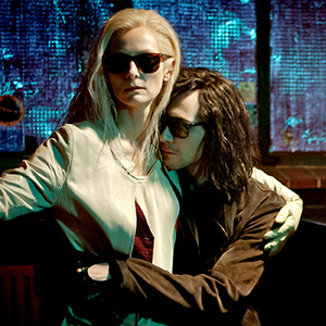 Two people with pale skin and long hair, both wearing dark sunglasses and stylish clothing, stand close together in a dimly lit room with glowing blue light from a window behind them. One person hugs the other protectively.
