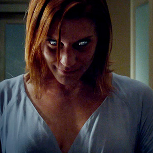 A person with shoulder-length red hair and glowing silver eyes stands indoors, wearing a light-colored shirt and looking menacingly downward with a sinister expression.
