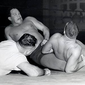 Black and white photo of two male wrestlers grappling on the mat as a referee closely observes the action, with one wrestler appearing to be in pain.