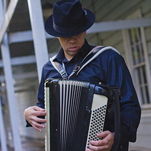 A person wearing a dark hat and shirt plays an accordion outdoors, with a building featuring pale blue siding and white railings in the background.