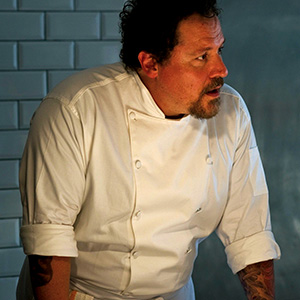 A chef wearing a white double-breasted jacket with rolled-up sleeves stands in front of a tiled wall, looking to the side with a focused expression.