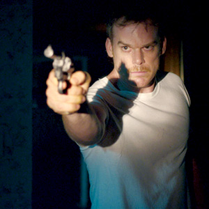A man in a white t-shirt points a revolver directly toward the camera with a serious expression, standing in a dimly lit room, partially illuminated by light.