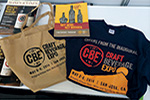 Expo merchandise including a Craft Beverage Expo tote bag, a flyer, and a navy blue t-shirt with event details and graphics are displayed on a table.