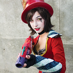 A person in a red costume with a matching top hat, dramatic makeup, and black gloves, points a colorful toy gun toward the camera against a plain concrete wall background.