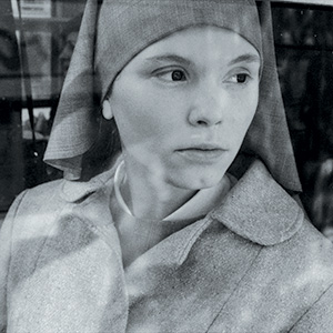 Black and white photo of a woman wearing a headscarf and coat, gazing pensively out of a window. Her expression is serious and contemplative, with soft light highlighting her face.