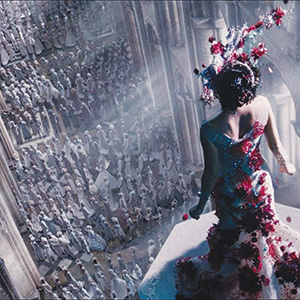 A woman in an elaborate gown covered with flowers stands on a balcony overlooking a large crowd of people dressed in white robes inside a grand, cathedral-like hall. Light streams down, creating a dramatic atmosphere.