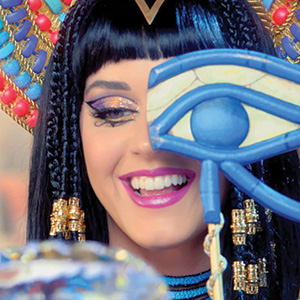 A woman with dark hair in Egyptian-style braids, colorful makeup, and a jeweled headdress smiles while holding a blue and gold Eye of Horus prop in front of her face.