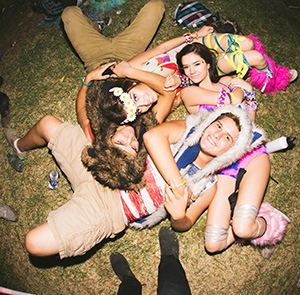 Four people wearing colorful, playful outfits and costumes lie on the grass in a close, happy group hug at an outdoor event or festival, smiling and enjoying themselves.