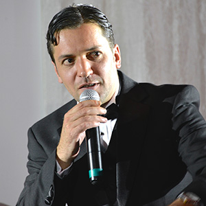 A man in a suit and bow tie is speaking into a microphone, looking slightly to the side, in a brightly lit indoor setting.