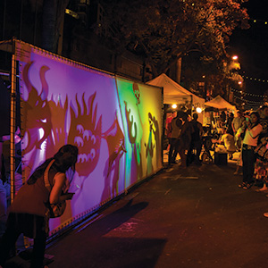 People watch colorful shadow puppets shaped like dragons and monsters projected on a screen at an outdoor night festival with tents and string lights in the background.