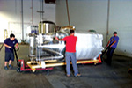 Three people use pallet jacks to move a large, shiny, industrial machine wrapped in protective material across a warehouse floor.