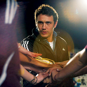 A man in a black jacket stands in a sports setting, placing his hand on top of a group huddle with several other people, suggesting teamwork and unity. Bright lights shine in the background.