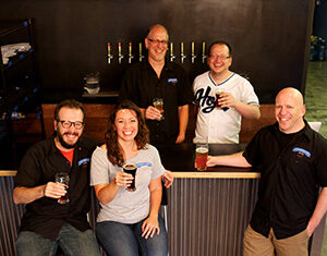 Five adults smiling and holding drinks inside a bar, with four standing behind the bar and one man and one woman sitting in front. Beer taps are visible in the background.