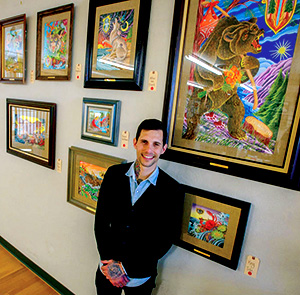 A person in a dark suit smiles while standing in front of a wall displaying several colorful, framed artworks, including a prominent painting of a bear playing a drum.