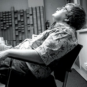 A person wearing glasses and a patterned shirt leans back in a chair, looking up with eyes closed in a room with shelves holding rows of cassette tapes or media in the background.