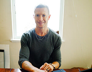 A man with short hair and a gray long-sleeve shirt sits at a wooden table, smiling at the camera while typing on a laptop. A bright window is in the background.
