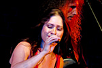 A woman sings passionately into a microphone on stage, with a guitarist featuring long curly hair playing in the background under stage lighting.