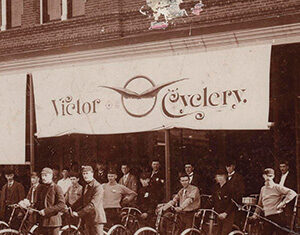 A vintage photo of a group of men standing with bicycles in front of a shop with a sign reading Victor Cyclery. The storefront has large windows and a brick exterior.