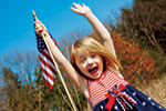 A young girl in a red, white, and blue dress smiles excitedly with both arms raised while holding an American flag outdoors, with trees and a blue sky in the background.