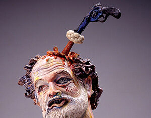 A colorful ceramic sculpture depicts a bearded mans severed head with a gun stuck in his skull, a knife in his neck, and a paintbrush in his shoulder, showing a surreal and dramatic expression.