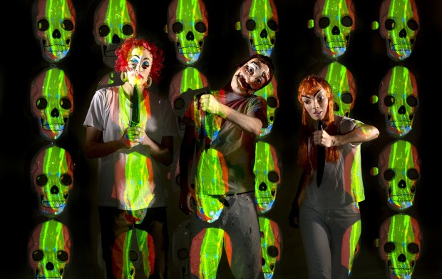 Three people in white clothes and creepy clown makeup pose holding knives, surrounded by a repeating pattern of neon green and red skulls on a dark background.