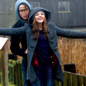 A young woman in a gray hooded coat stands with her arms outstretched, smiling, while a young man in a blue hoodie stands closely behind her. They are outdoors on a cloudy day.