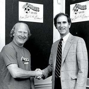 Two men smiling and shaking hands in front of signs that read Quakes San Jose with a soccer ball logo. One man wears a t-shirt; the other wears a suit and tie. Black and white photo.