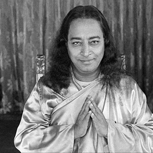 A man with long hair and a serene smile sits with his hands pressed together in a gesture of greeting or prayer, wearing a light-colored robe, against a softly patterned curtain background.