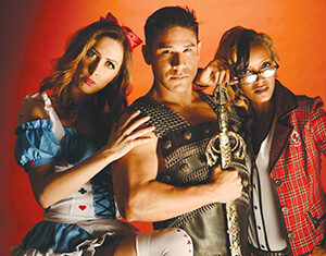 Three people in costumes pose against a red background: a woman in an Alice in Wonderland outfit, a man in medieval armor holding a sword, and a woman in a schoolgirl outfit and glasses.