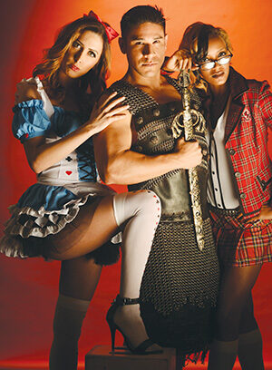 Three people in costumes pose against a red background: a woman in an Alice in Wonderland outfit, a man in medieval armor holding a sword, and a woman in a schoolgirl outfit and glasses.