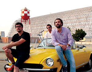 Three men sit on and around a yellow convertible car parked outdoors. A large hangar and a red and white water tower are visible in the background. The scene appears relaxed and casual.