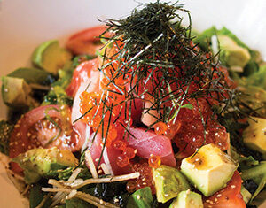 A colorful salad with diced avocado, mixed greens, seaweed strips, salmon roe, sliced fish, and tomatoes, garnished with sesame seeds and thin julienned vegetables.