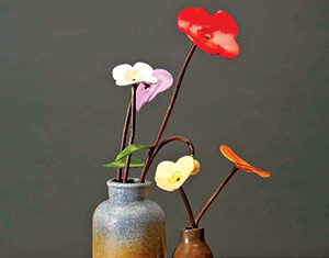 Two ceramic vases with colorful, artificial flowers in red, white, purple, yellow, and orange, arranged on a wooden base against a plain dark background.