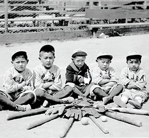 Five young boys in baseball uniforms sit cross-legged in a semicircle on a dirt field with bats, gloves, and balls arranged in front of them. Empty bleachers are visible in the background.