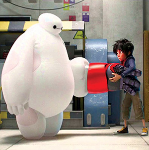 A large white inflatable robot extends its arm while a boy with dark hair, wearing a jacket and shorts, attaches a red mechanical hand to the robot. They are in a workshop setting.