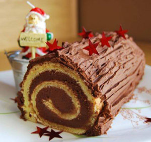 A chocolate yule log cake with a swirl pattern, decorated with red star-shaped sprinkles. A small Santa figurine holding a Welcome sign stands in the background.