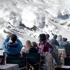 People sitting at outdoor restaurant tables on a snowy mountain watch an avalanche cascading down the slopes in the distance. Some look alarmed, turning to each other, while others watch the scene unfold.
