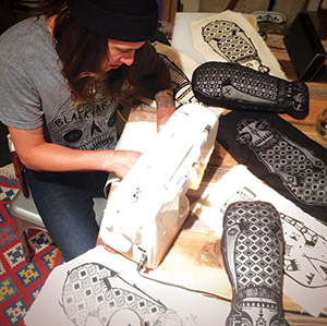 A person wearing a beanie uses a sewing machine at a table covered with fabric pieces featuring intricate black and white patterns, including skateboards and geometric designs.