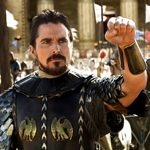 A man in ancient warrior armor with decorative eagle emblems raises his fist. He has short dark hair, a beard, and stands in front of grand stone columns, suggesting a historical or epic setting.