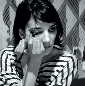 A woman with short dark hair and a striped shirt applies eyeliner while looking in a mirror, with a geometric-patterned background behind her. The image is in black and white.
