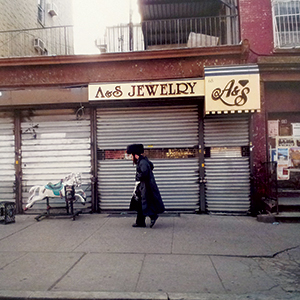A person in dark clothing and a fur hat walks past a closed jewelry store with metal shutters and an old sign reading A&S Jewelry. A small white carousel horse is on the sidewalk nearby.