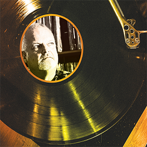 A vinyl record on a turntable; the center label features a circular photo of an older man with a white beard and mustache, looking up thoughtfully, with shelves of records in the background.